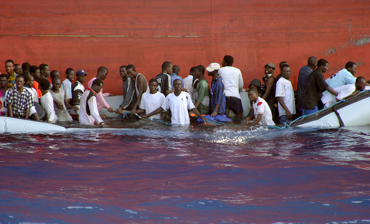 A group of migrants in a half-submerged dinghy await rescue by the Armed Forces of Malta while sheltering against the hull of a cargo ship.
